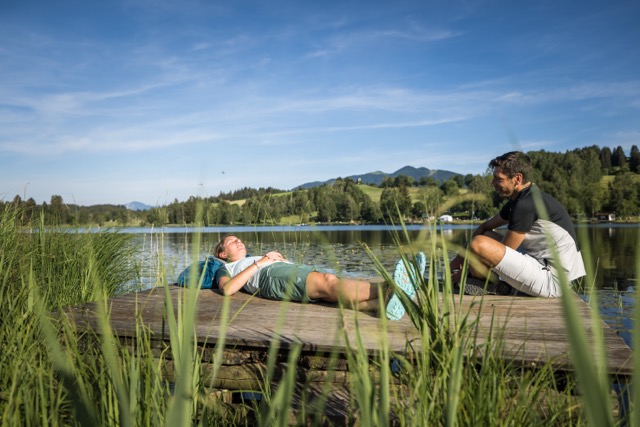 Wanderer liegen auf einem Steg, im Hintergrund ein See und die Bergkulisse der Ammergauer Alpen