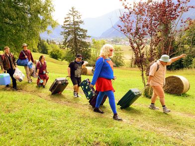Six people with luggage and suitcases on a meadow, with trees and mountains in the background.