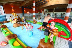 Kinder spielen im Familienbecken eines Hallenbads mit Wasserspielgerät und kleiner Rutsche.