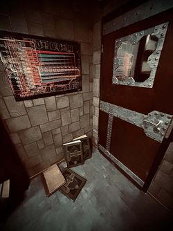 Dunkler Escape-Room mit technischem Wandplan, schwerer Metalltür und Büchern als Rätselrequisiten.