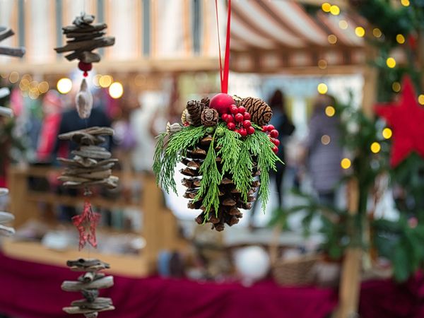 Es ist ein geschmückter Tannenzapfen vor einem beleuchteten Christlkindlmarktstand zu sehen. 