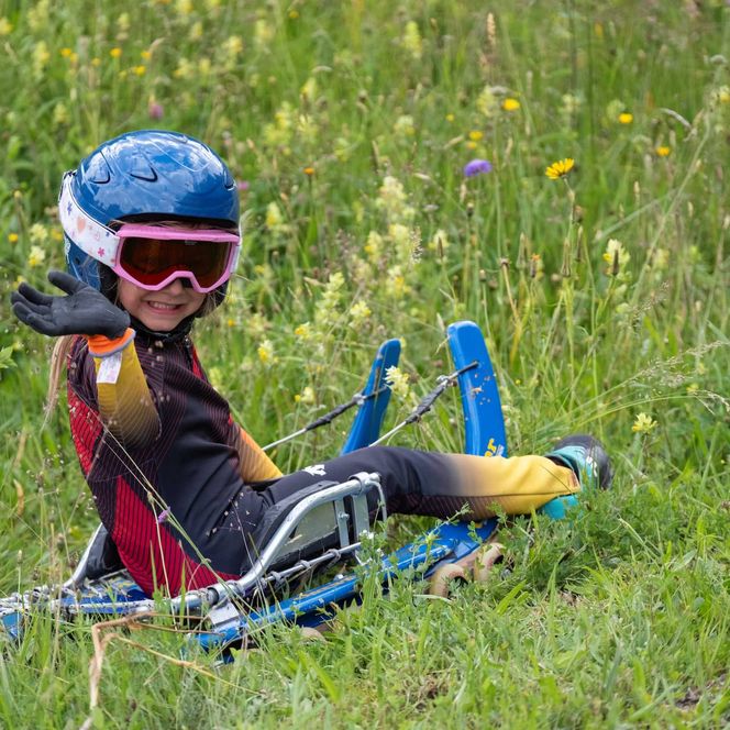 Kind mit Helm und Schutzbrille sitzt auf einem Rollenrodelschlitten in einer Blumenwiese und winkt.