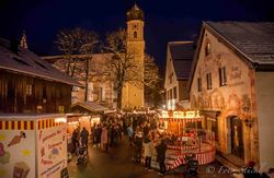 Der Christkindlmarkt im winterlichen Oberammergau mithell erleuchteter Pfarrkirche und mit Lüftlmalereien verzierte Häuser.