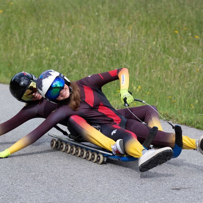 Zwei Personen fahren im Doppelsitzer auf einem Rollenrodel durch eine Kurve auf Asphalt.